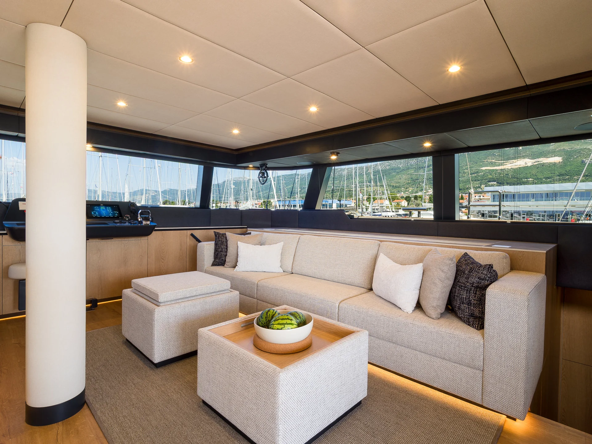 Interior of a yacht cabin featuring a light-colored sofa, an ottoman, and a square coffee table topped with a bowl of watermelons, with large windows overlooking a marina.