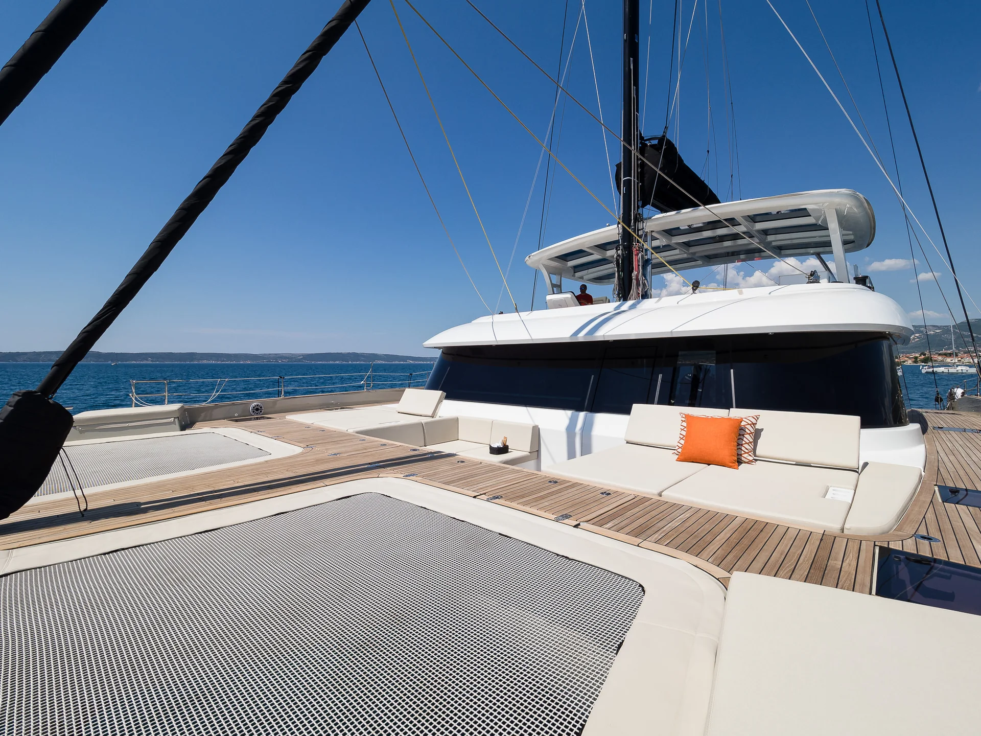 The deck of a catamaran featuring a mesh trampoline and white cushioned seating with an orange throw pillow under a clear blue sky.