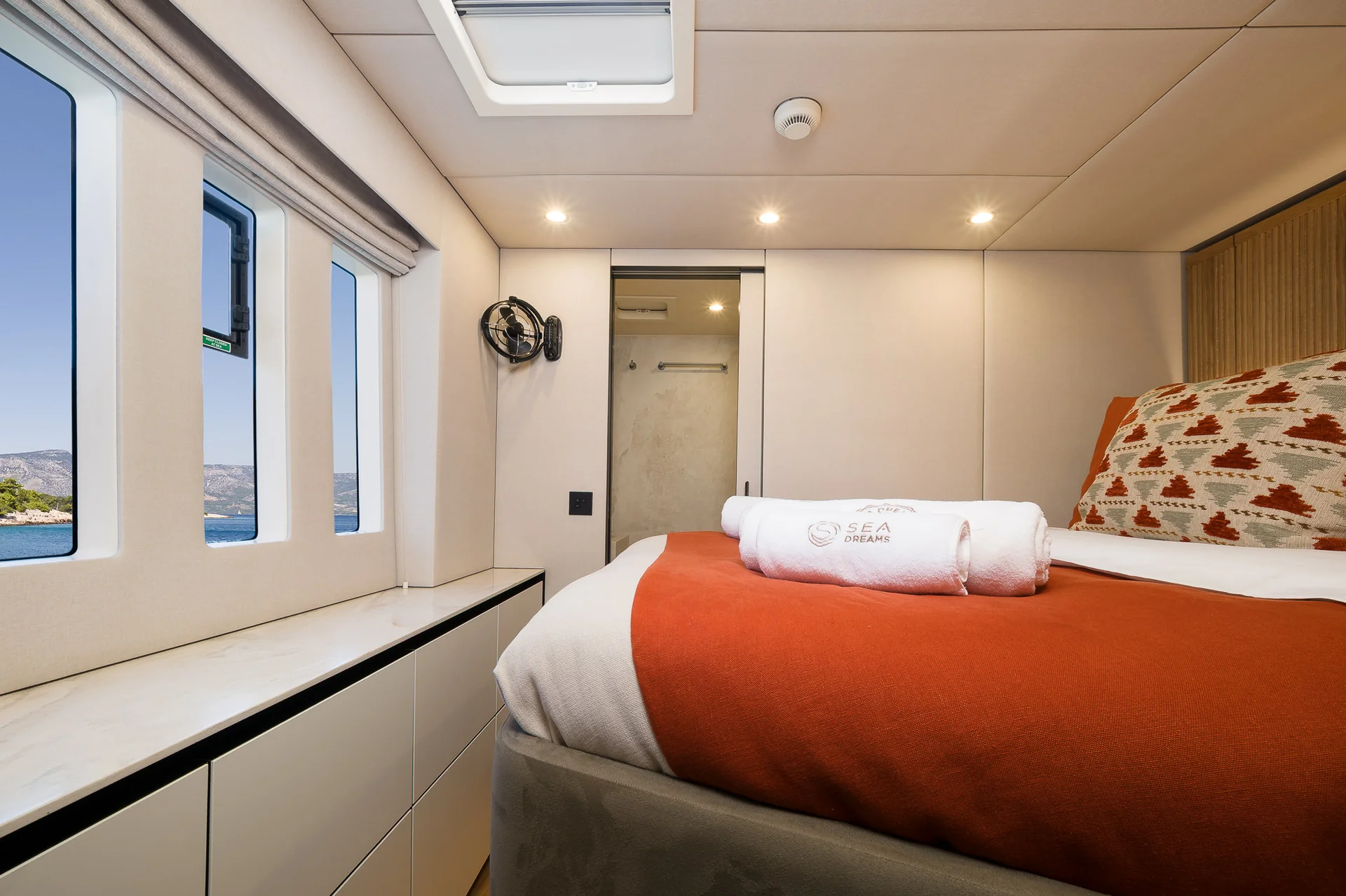 A bed with a red runner and patterned pillows in a yacht cabin, featuring two rolled towels branded Sea Dreams and a view of the sea through the windows.