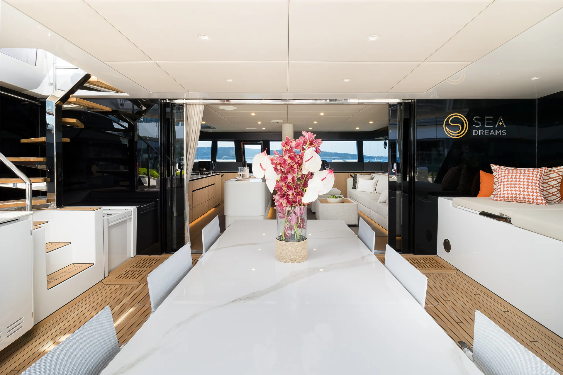A marble dining table with a large vase of orchids on the aft deck of a yacht, overlooking an interior lounge and the sea.