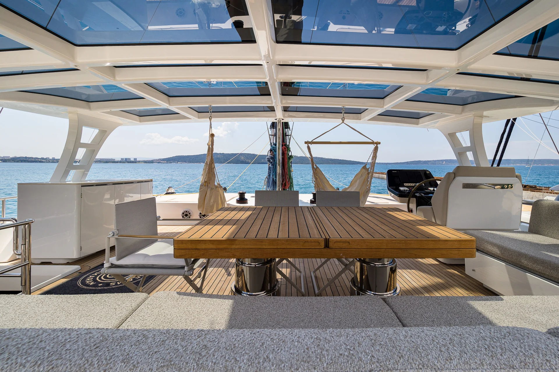 The outdoor deck of a catamaran with a wooden table, grey chairs, and two hanging chairs, overlooking the open sea.