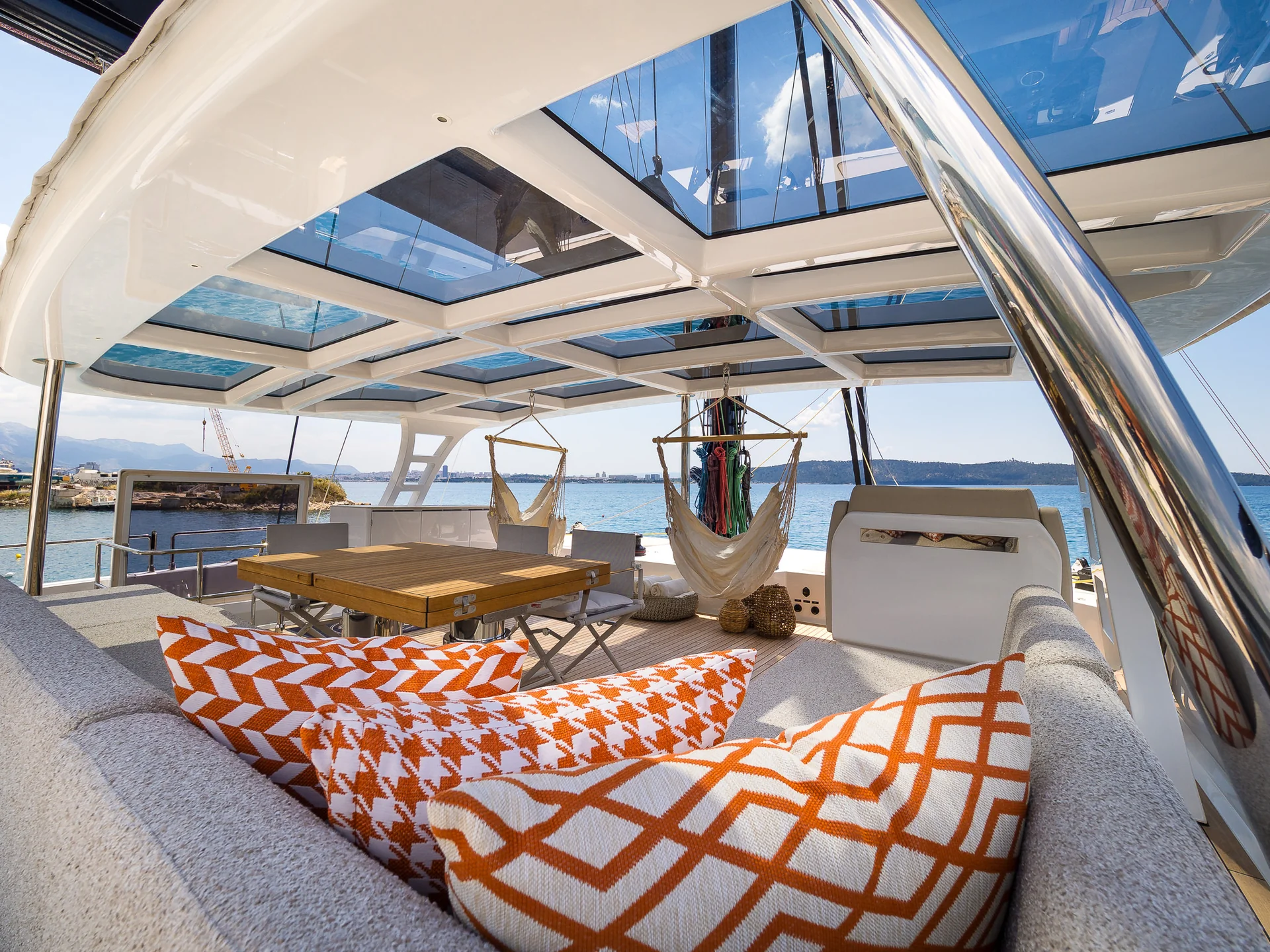 A view from a lounge area on a boat deck with orange-patterned throw pillows, a wooden dining table, and two hanging chairs under a glass ceiling overlooking the ocean.